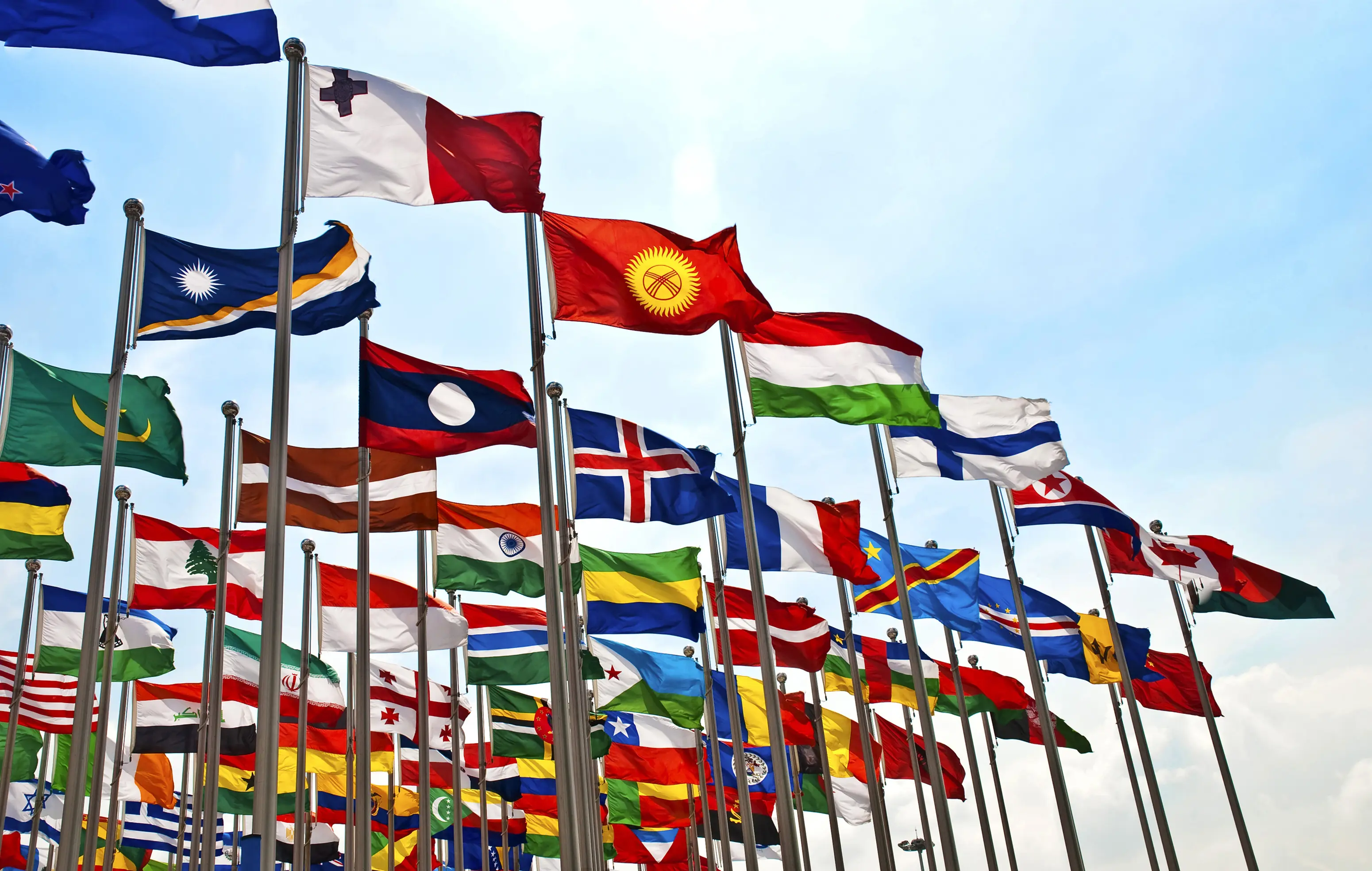Multiple international flags waving on flagpoles against a clear sky.