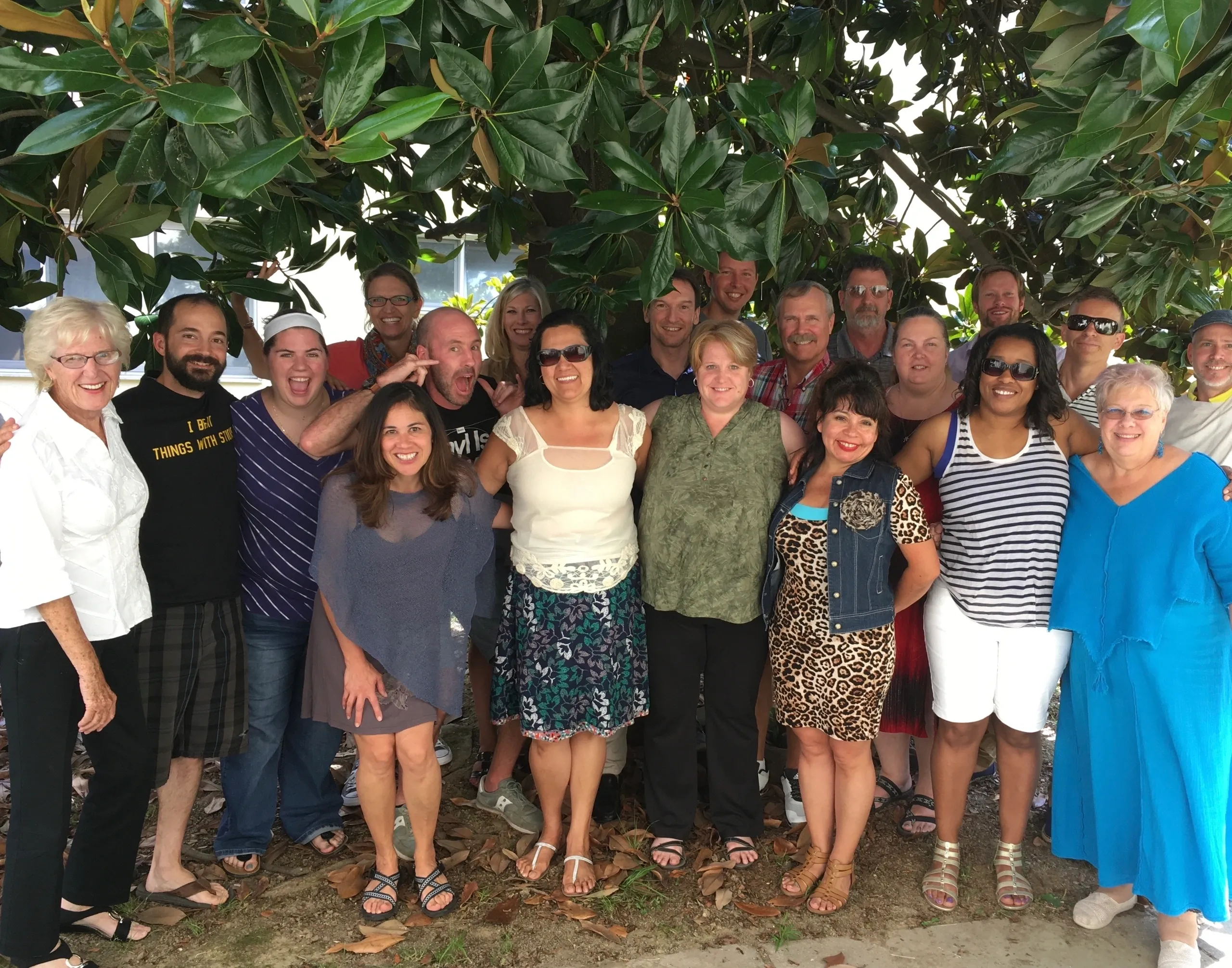 Group photo of people smiling under a leafy tree outdoors.